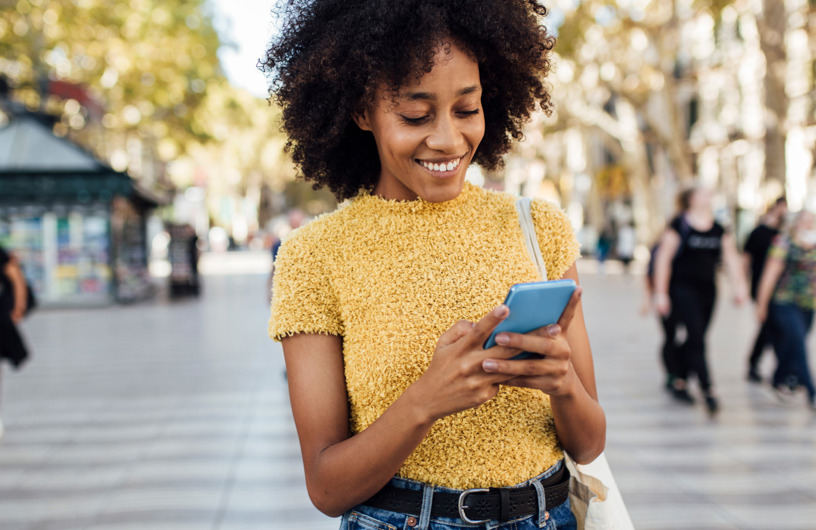 Ung kvinde iført gul bluse og jeans, står midt på et torv og smiler ad hendes telefon. 